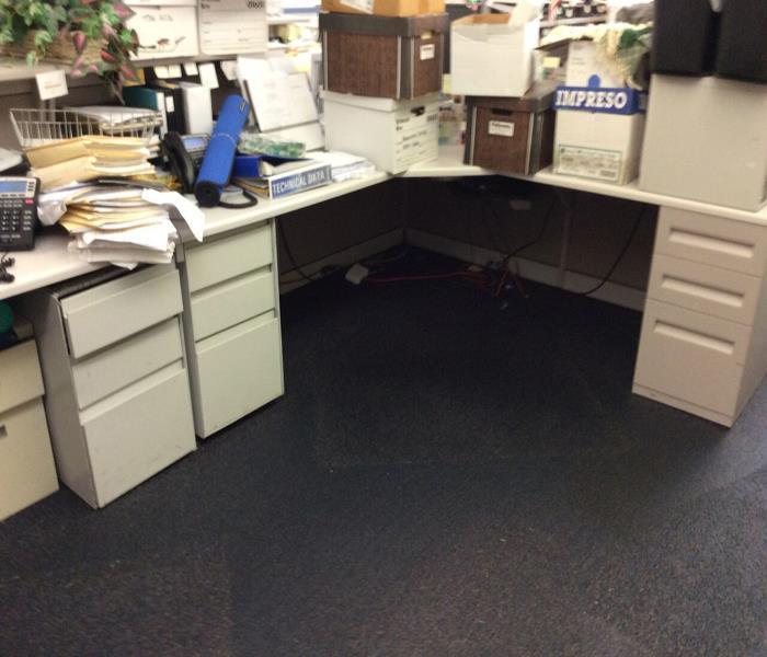 Image shows cubicle desk with water underneath on the carpet, and files and boxes piled on the desk out of the way.